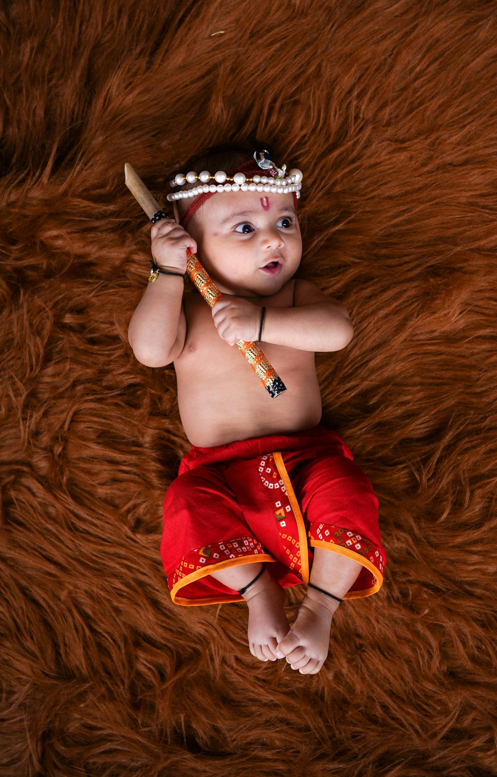 Cute baby dressed in traditional attire holding a stick, lying on a soft brown surface.